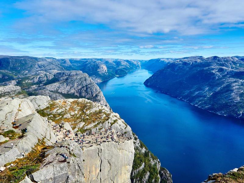Hikers on a flat cliff ledge overlooking a deep blue fjord and mountain landscape